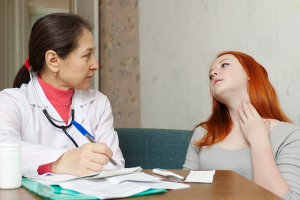 A woman physician takes notes as a patient tilts her head and points to her throat. 