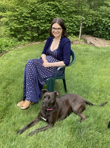 Erica, wearing glasses and a navy blue patterned dress, smiling and seated on a green plastic chair on a grassy lawn. Next to her, a black dog with a pink collar rests on the ground, with greenery in the background.