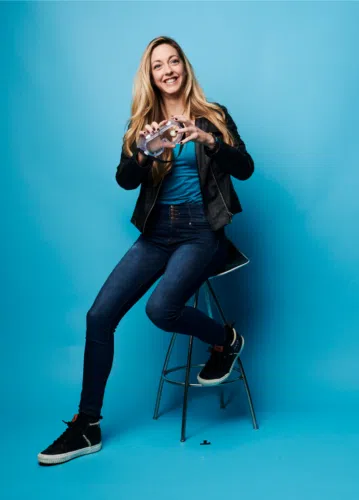 Dianna Cowern sitting on a high stool against a solid blue background. She has long blond hair and is smiling, giving a joyful and relaxed vibe.