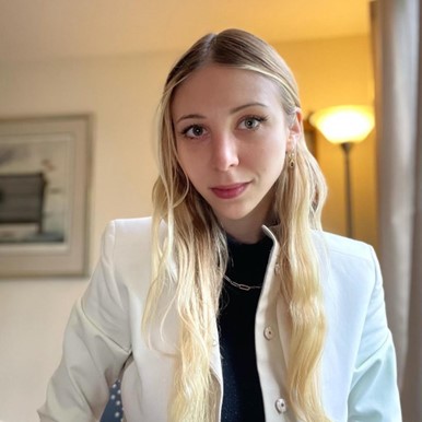 Picture of Rebecca with long, light hair, wearing a white jacket over a dark top. She is indoors, and there is a floor lamp and a framed picture visible in the background, contributing to a cozy and softly lit atmosphere.