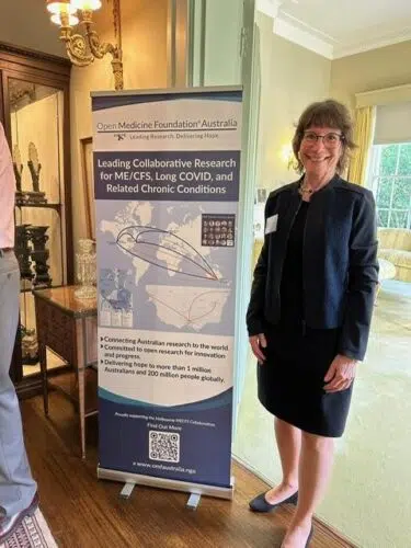 OMF Founder and CEO Linda Tannenbaum stands indoors next to a vertical banner for Open Medicine Foundation Australia. She is smiling and dressed in a black suit with a matching skirt and jacket.