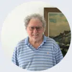 Keith is standing indoors in front of a white wall with a framed painting in the background. He has curly gray hair, a short white beard, and is wearing glasses.