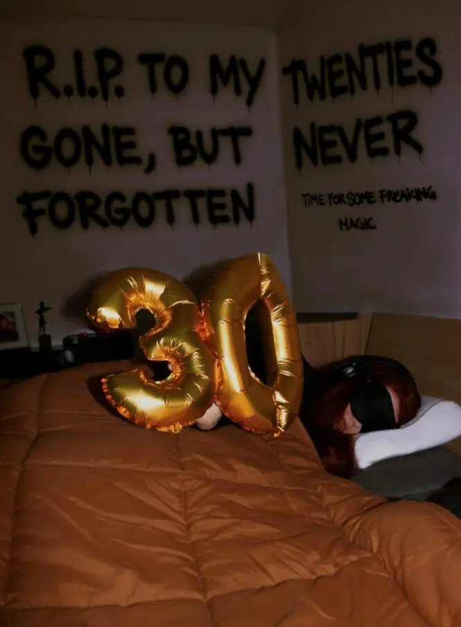 In a dark room, golden foil balloons shaped as the number "30" rest on top of a brown comforter. Behind the balloons, Bente—a person living with very severe ME—is lying in bed, wearing a sleep mask and noise-canceling headphones.The wall behind her features bold, black text that reads: 'R.I.P. TO MY TWENTIES — GONE, BUT NEVER FORGOTTEN’.