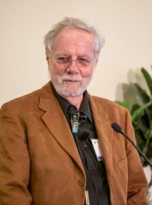 Photo of Dr. Ron Davis standing in front of the microphone. He wears a warm brown blazer over a black shirt.