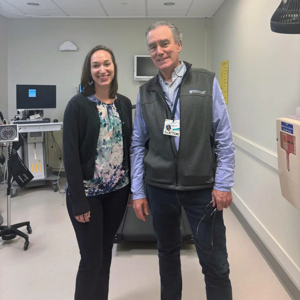 Dr. Meadows and Dr. Systrom stand smiling in a hospital room. Dr. Meadows wears a floral blouse and black cardigan; Dr. Systrom wears a gray vest with a badge labeled “Doctor”.