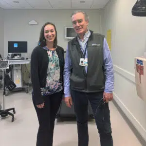 Dr. Meadows and Dr. Systrom stand smiling in a hospital room. Dr. Meadows wears a floral blouse and black cardigan; Dr. Systrom wears a gray vest with a badge labeled “Doctor”.