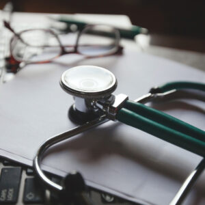 Close-up of a stethoscope resting on a stack of papers with a pair of eyeglasses in the background, suggesting a clinical setting.