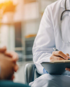 Close-up of a doctor in a white coat with a stethoscope writing on a clipboard while listening to a patient during a clinic visit.